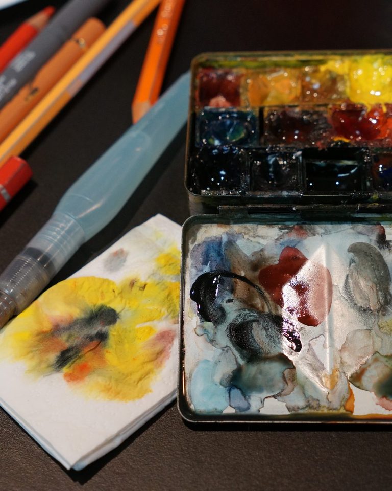 Watercolor palette, water brush, and colored pencils on a dark table